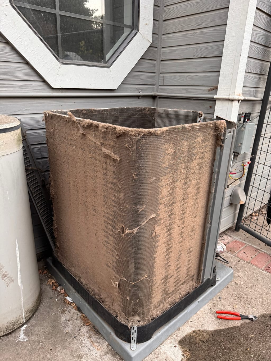 Dust and lint covering the Rheem outdoor coil below the octagonal window before cleaning in Clayton